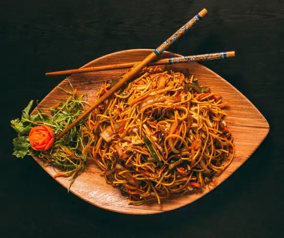   A top-down shot of a wooden, leaf-shaped plate filled with a large serving of noodles, garnished with a side of green leafy vegetables and an intricately carved orange garnish, with a pair of chopsticks resting on the plate.