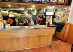 A wooden service counter in a restaurant, with a glimpse into the kitchen showing stainless steel equipment, a refrigerator, and a person in a red shirt.