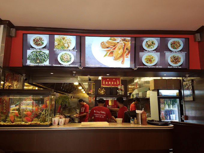 Interior of Wa Lung Kitchen with staff preparing food, a fish tank at the counter, and digital menu screens displaying various Chinese dishes