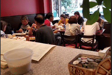 A group of people are seated at tables inside a restaurant, with a counter and a tip jar visible in the foreground.