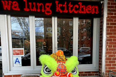 Close-up of the Wa Lung Kitchen sign with red lettering above a window, with a decorative lion dance head in the foreground.