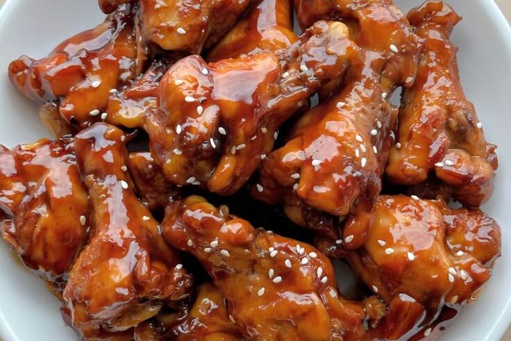 A plate of glistening, honey-glazed chicken wings sprinkled with white sesame seeds, showcasing their crispy texture and rich color.
