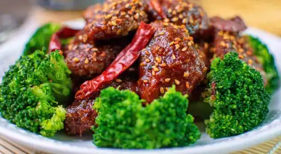 Spicy Garlic Beef with Broccoli and Dried Red Chilies A white plate featuring spicy garlic-coated beef chunks topped with crushed red pepper and garnished with whole dried red chilies, surrounded by steamed vibrant green broccoli florets.
