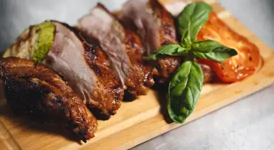 Grilled Sliced Pork with Roasted Tomato and Basil A wooden serving board with thick slices of grilled pork, charred on the outside and juicy inside, accompanied by a roasted tomato slice, grilled zucchini, and fresh basil leaves as garnish.