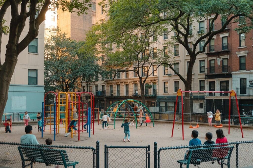 Children playing on colorful playground equipment surrounded by benches and tall city buildings at a small urban park on a sunny afternoon.